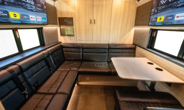 Daylit rear lounge of the Astro celebrity coach with U-shaped leather seating, table, side windows and dual smart TVs